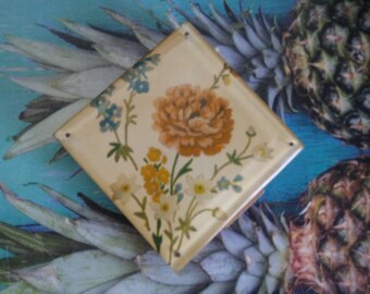 May include: A square, cream-colored tile featuring a central orange flower surrounded by smaller yellow and blue floral illustrations. The tile has a smooth, glossy finish and small holes at the corners, displaying a botanical design on its surface.