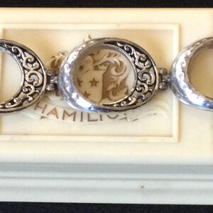 Może przedstawiać: Srebrna bransoletka z otwartymi owalnymi ogniwami i ozdobnymi zawijasami. Bransoletka jest prezentowana na kremowym pudełku z kwiatowym wzorem i napisem "HAMILTON".