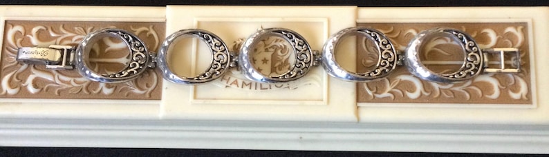 Może przedstawiać: Srebrna bransoletka z otwartymi owalnymi ogniwami i ozdobnymi zawijasami. Bransoletka jest prezentowana na kremowym pudełku z kwiatowym wzorem i napisem "HAMILTON".