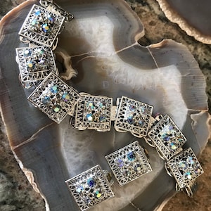 Pu&ograve; includere: Un set di braccialetto e orecchini in tono argento con strass iridescenti a forma quadrata. I gioielli sono in metallo con un design a filigrana.