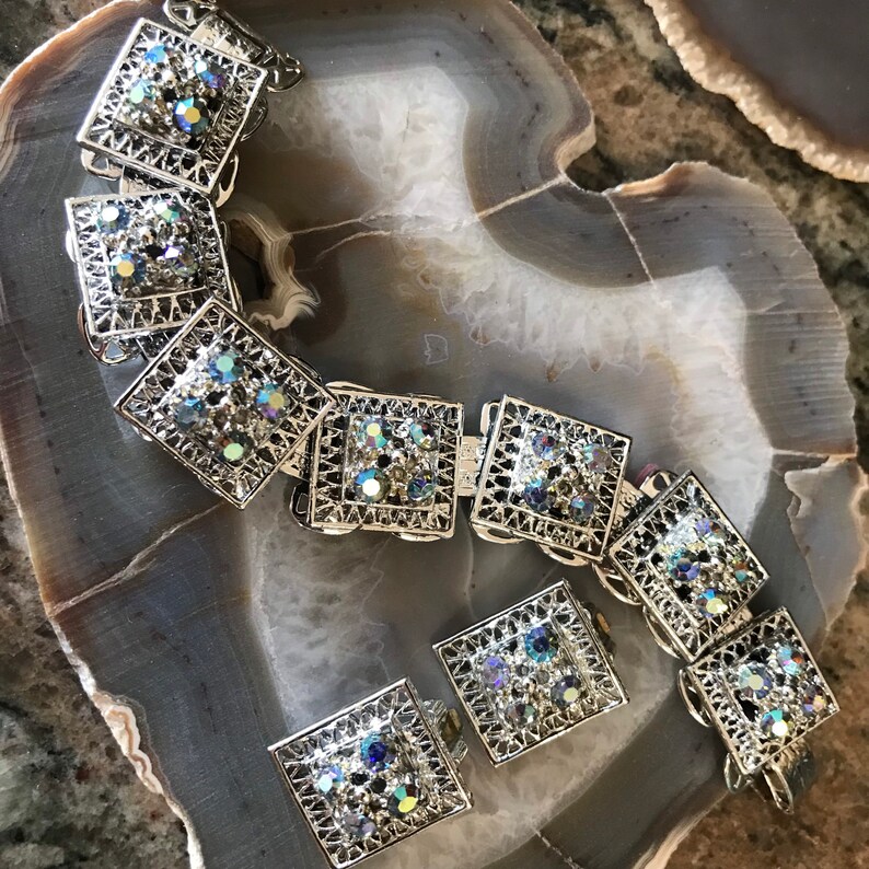 Pu&ograve; includere: Un set di braccialetto e orecchini in tono argento con strass iridescenti a forma quadrata. I gioielli sono in metallo con un design a filigrana.