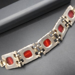 May include: A silver-toned bracelet with rectangular red stones. The bracelet is composed of connected rectangular links, each housing a red stone. The clasp is visible at the end of the bracelet. The bracelet is on a dark surface.