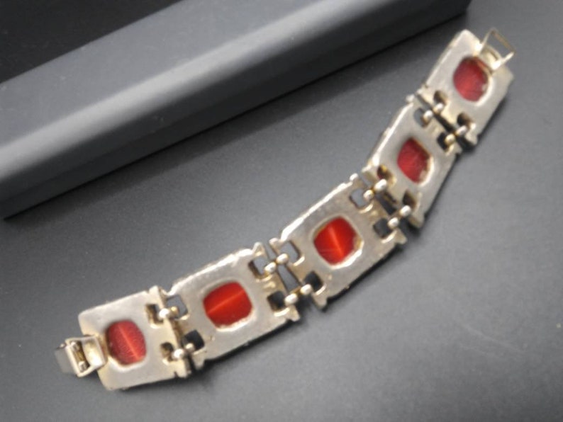 May include: A silver-toned bracelet with rectangular red stones. The bracelet is composed of connected rectangular links, each housing a red stone. The clasp is visible at the end of the bracelet. The bracelet is on a dark surface.