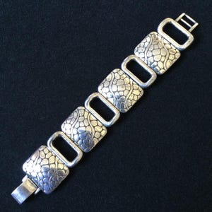 May include: A silver-toned link bracelet featuring four square-shaped segments with a raised, pebble-like texture alternating with four smooth, open rectangular links. The bracelet includes a metal clasp mechanism at one end for secure fastening.