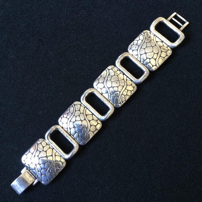 May include: A silver-toned link bracelet featuring four square-shaped segments with a raised, pebble-like texture alternating with four smooth, open rectangular links. The bracelet includes a metal clasp mechanism at one end for secure fastening.