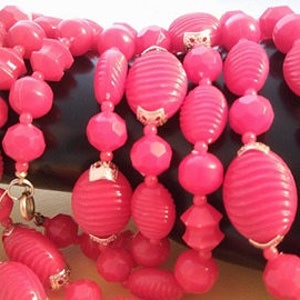 May include: A multi-strand necklace made of pink plastic beads. The beads are of various shapes and sizes, including round, oval, and faceted. The necklace is strung on a gold chain.