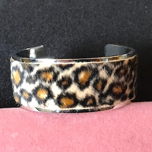 May include: A wide cuff bracelet with a silver-tone metal frame. The center band features a leopard print pattern in shades of brown, black, and cream. The bracelet has a polished finish and a modern design.