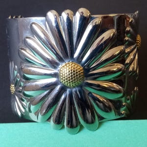 Könnte beinhalten: Silberfarbenes Armreif-Armband mit einem Gänseblümchen-Design. Das breite Band hat erhabene Gänseblümchen-Blütenblätter und eine strukturierte goldfarbene Mitte. Das Blumenmuster wiederholt sich um das Armband.
