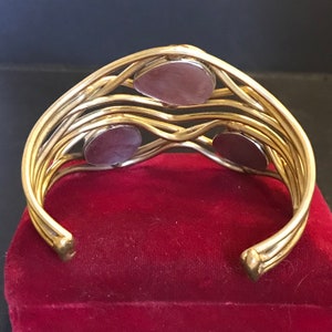 May include: Gold tone bangle bracelet with three round, dark purple stones set into the metal. The bracelet is open and has a woven design.