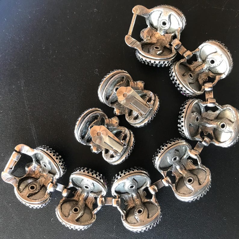 Peut inclure: Boucles d'oreilles &agrave; clip et bracelet assorti de couleur argent&eacute;e vintage. Les bijoux pr&eacute;sentent un design d&eacute;taill&eacute; avec de petits accents sombres sur les bords. Les pi&egrave;ces sont dispos&eacute;es sur une surface sombre, mettant en valeur leur savoir-faire.