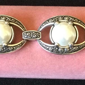 Peut inclure: Bracelet argent&eacute; compos&eacute; de quatre maillons ovales, chacun orn&eacute; d'une perle blanche centrale. Les maillons sont reli&eacute;s par de petites sections d&eacute;coratives. Le bracelet est pos&eacute; sur un fond rose.