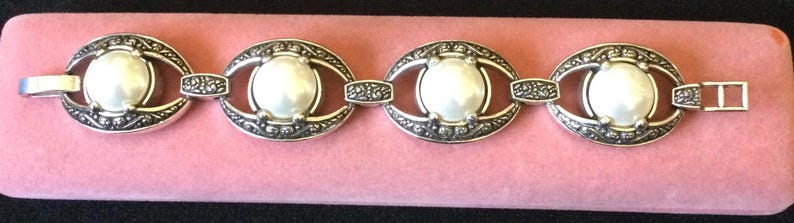 Peut inclure: Bracelet argent&eacute; compos&eacute; de quatre maillons ovales, chacun orn&eacute; d'une perle blanche centrale. Les maillons sont reli&eacute;s par de petites sections d&eacute;coratives. Le bracelet est pos&eacute; sur un fond rose.