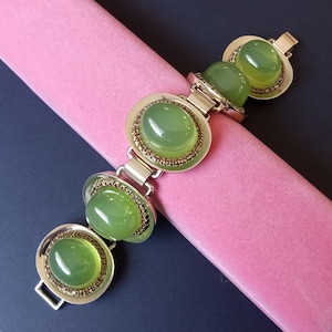 Könnte beinhalten: Ein goldenes Armband mit vier großen, ovalen, grünen Steinen. Jeder Stein ist in einer goldenen Fassung mit einem dekorativen Rand gefasst.