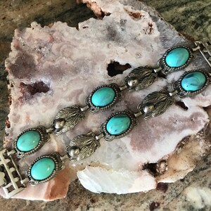 Op de afbeelding: Twee zilverkleurige armbanden met ovale, turquoise stenen. De armbanden hebben een decoratief blad- en kralenontwerp. De sieraden worden gepresenteerd op een lichtroze en wit stenen oppervlak.