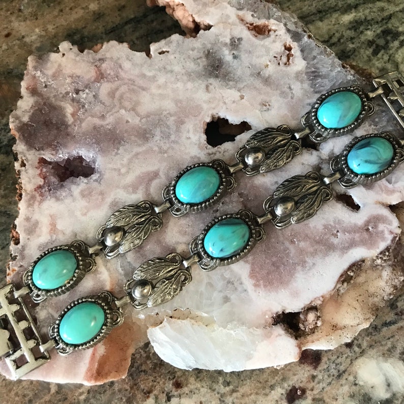 Op de afbeelding: Twee zilverkleurige armbanden met ovale, turquoise stenen. De armbanden hebben een decoratief blad- en kralenontwerp. De sieraden worden gepresenteerd op een lichtroze en wit stenen oppervlak.