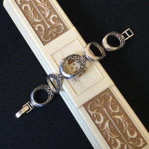 Może przedstawiać: Srebrna bransoletka z owalnymi ogniwami i skomplikowanymi ażurowymi wzorami. Bransoletka jest prezentowana na kremowym pudełku z dekoracyjnym wzorem kwiatowym. Zapięcie jest widoczne, a bransoletka wydaje się być wykonana z metalu.