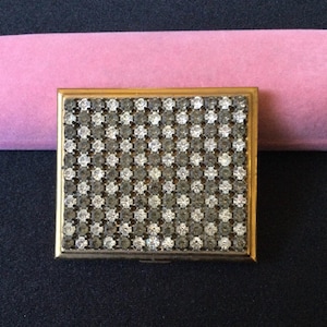 May include: A rectangular, gold-toned compact mirror with a surface covered in a grid pattern of clear and smoky-gray rhinestones. The compact is set against a black surface with a pink fabric in the background.