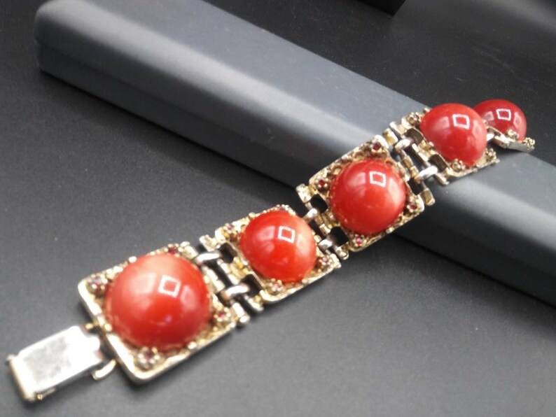 May include: A vintage bracelet featuring large, round, red cabochon stones set in gold-toned metal frames. The bracelet has a silver-colored clasp and is accented with small, reddish-brown stones. The design is geometric and classic.