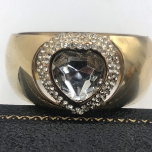 Puede incluir: Un brazalete dorado con un gran cristal transparente en forma de coraz&oacute;n rodeado de peque&ntilde;os diamantes de imitaci&oacute;n.