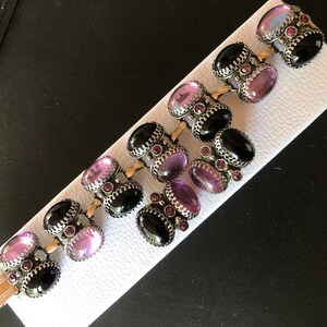 Peut inclure: Un bracelet vintage orn&eacute; de pierres pr&eacute;cieuses ovales noires, violettes et roses. Les pierres sont serties dans des montures en m&eacute;tal argent&eacute; orn&eacute;es de petites pierres rouges. Le bracelet est pr&eacute;sent&eacute; sur une surface blanche textur&eacute;e.