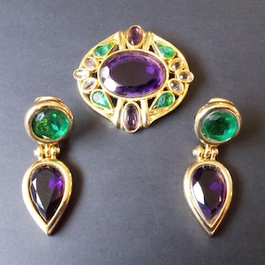 Peut inclure: Une broche et des boucles d'oreilles assorties en or avec des pierres pr&eacute;cieuses vertes et violettes. La broche pr&eacute;sente une grande pierre violette de forme ovale entour&eacute;e de pierres vertes et claires plus petites. Les boucles d'oreilles pr&eacute;sentent une pierre ronde verte et une pierre violette en forme de larme.