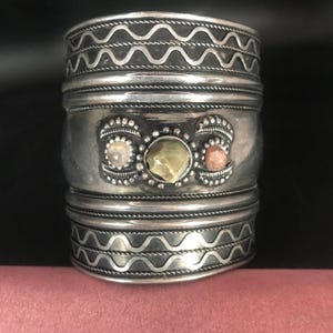Könnte beinhalten: Ein breites, silberfarbenes Armreif-Armband. Das Armband hat dekorative Bänder mit einem wellenförmigen Design. In der Mitte des Armbands befinden sich drei eingesetzte Steine: ein hellrosa, ein grüner und ein rot-oranger Stein. Ein auffälliges Schmuckstück.
