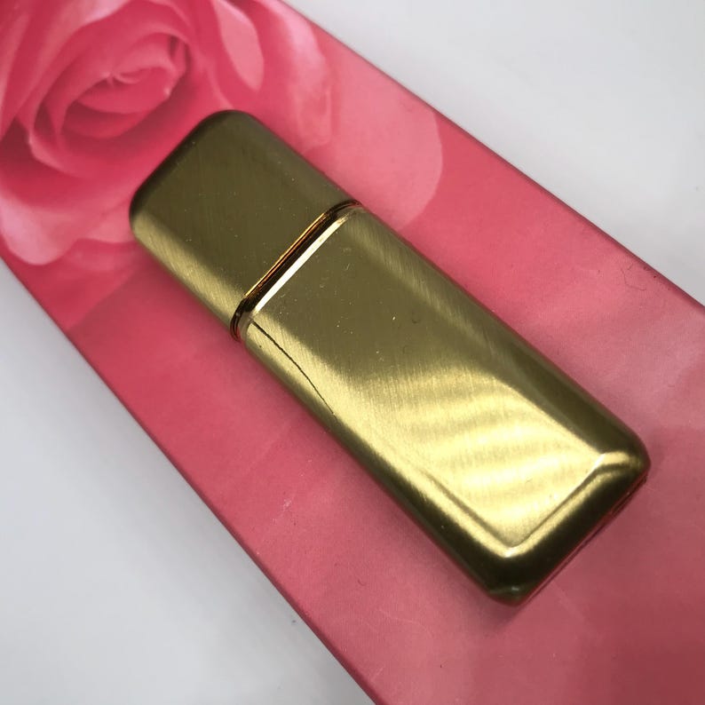 May include: A gold metal perfume bottle with a brushed finish. The bottle is rectangular with rounded edges and has a thin gold band around the middle.