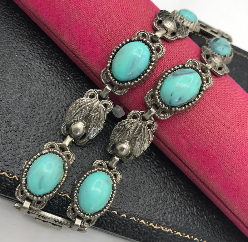 Op de afbeelding: Vintage zilverkleurige armband met ovale turquoise stenen. De armband heeft een sierlijk ontwerp met bladvormige accenten en een veilige sluiting. De stenen hebben een gemarmerd uiterlijk.