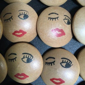 May include: Close-up of several wooden, oval-shaped faces with painted features. Each face has closed eyes, black eyelashes, and red lips. The wooden faces are a light brown color, and the painted features are black and red.