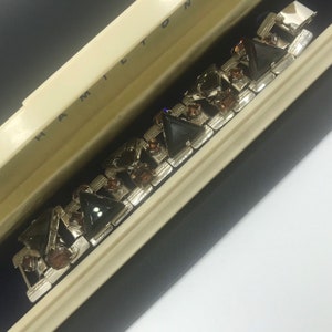 May include: A silver bracelet with black and brown triangular stones in a white box with the word "HAMILTON" printed on the lid.