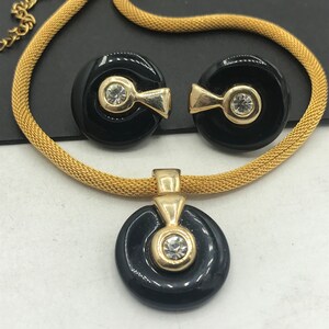 May include: A gold chain necklace with a black pendant featuring a gold circle with a clear rhinestone. Matching earrings with black circles and gold accents with clear rhinestones.