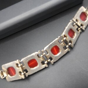 May include: A silver-toned bracelet with rectangular red stones. The bracelet is composed of connected rectangular links, each housing a red stone. The clasp is visible at the end of the bracelet. The bracelet is on a dark surface.