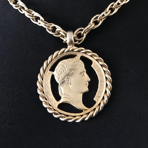 May include: Gold-toned necklace featuring a circular pendant with a profile of a person wearing a laurel wreath. The pendant has a rope-like border and is suspended from a matching chain. The background is black.