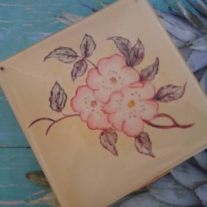 May include: A square, cream-colored ceramic tile featuring a hand-painted floral design. The illustration depicts three soft pink blossoms with yellow centers, surrounded by dark grey-toned leaves and stems. Two small metal pins are visible at the corners.