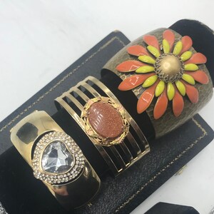Puede incluir: Tres pulseras doradas con diferentes adornos. La primera pulsera tiene un gran dise&ntilde;o de flor naranja y amarilla. La segunda pulsera tiene una piedra marr&oacute;n engastada en un marco dorado. La tercera pulsera tiene un coraz&oacute;n de cristal transparente.