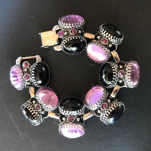 Peut inclure: Un bracelet argent&eacute; orn&eacute; de pierres ovales noires et lavande. Le bracelet pr&eacute;sente un design d&eacute;coratif avec de petites pierres roses et une fermeture &agrave; fermoir. Le bracelet est pr&eacute;sent&eacute; sur une surface sombre.
