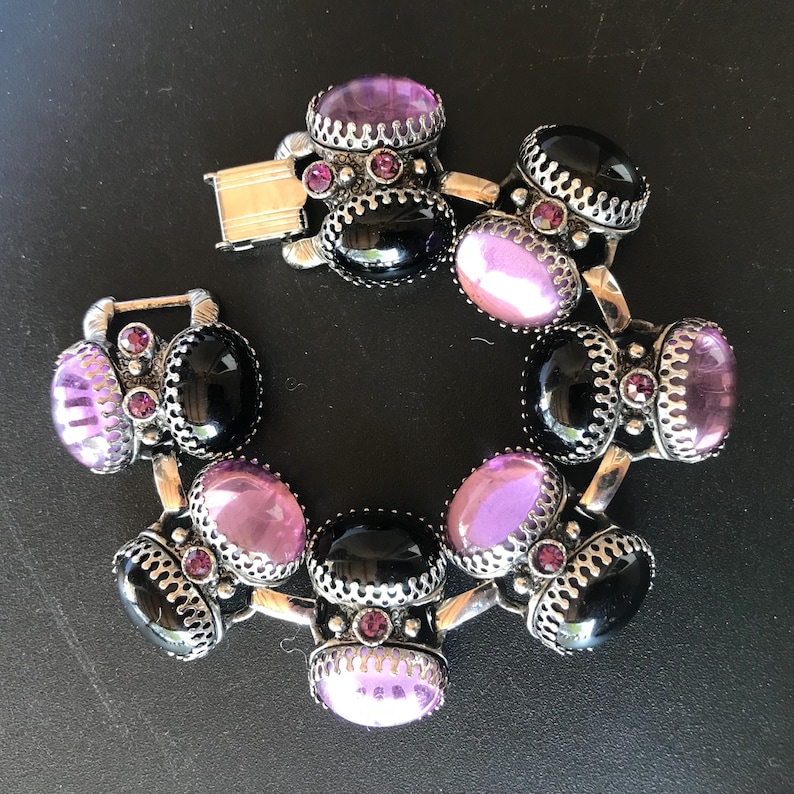 Peut inclure: Un bracelet argent&eacute; orn&eacute; de pierres ovales noires et lavande. Le bracelet pr&eacute;sente un design d&eacute;coratif avec de petites pierres roses et une fermeture &agrave; fermoir. Le bracelet est pr&eacute;sent&eacute; sur une surface sombre.