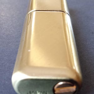 May include: A gold-colored metal lighter with a rounded rectangular shape. The lighter has a small, round, copper-colored button on the side and the word "Japan" engraved on the bottom.