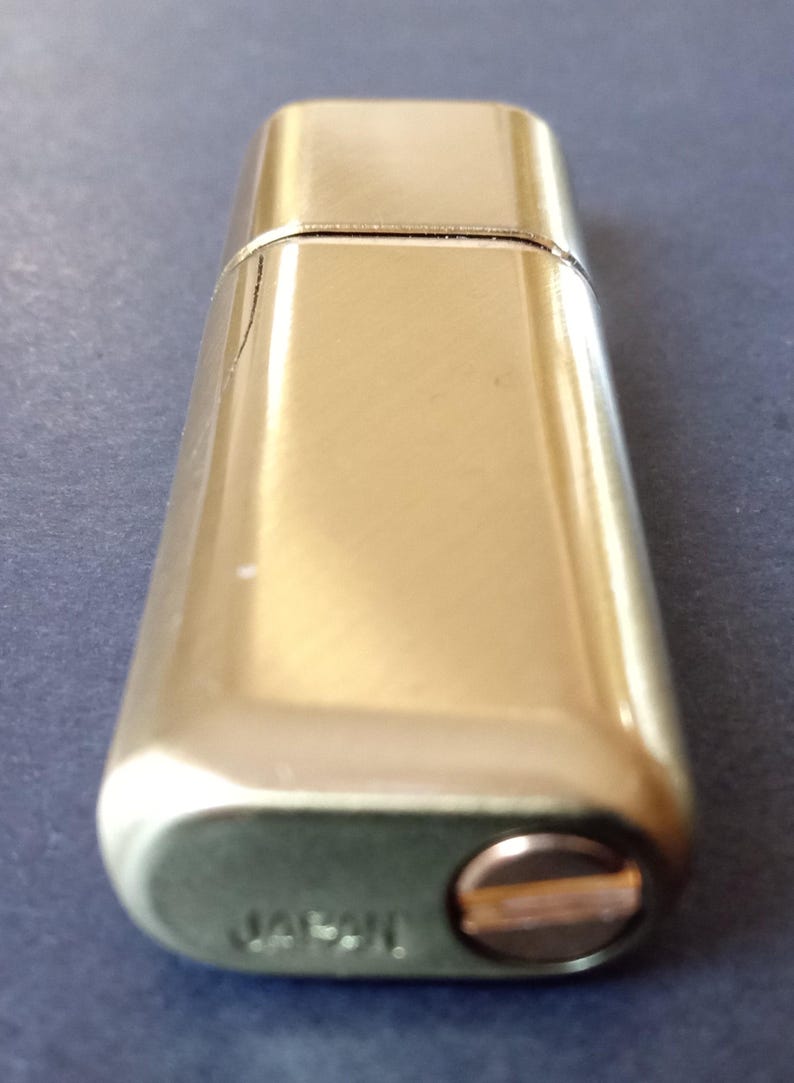 May include: A gold-colored metal lighter with a rounded rectangular shape. The lighter has a small, round, copper-colored button on the side and the word "Japan" engraved on the bottom.