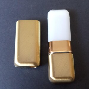 May include: Two gold-colored metal lipstick cases, one with a white top and one without. The cases have a brushed finish.