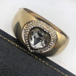 Puede incluir: Un brazalete de oro con un gran cristal transparente en forma de coraz&oacute;n rodeado de cristales m&aacute;s peque&ntilde;os.