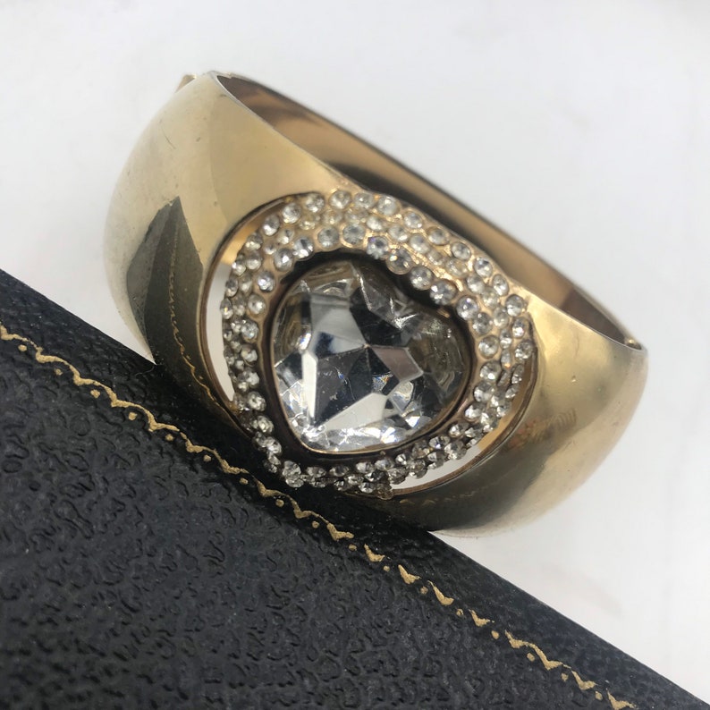Puede incluir: Un brazalete de oro con un gran cristal transparente en forma de coraz&oacute;n rodeado de cristales m&aacute;s peque&ntilde;os.