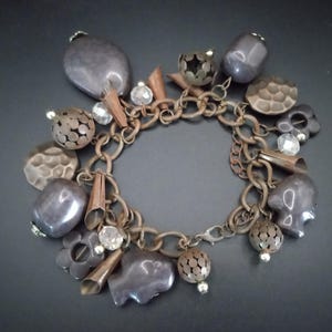 May include: A silver chain bracelet with multiple charms, including a variety of metal shapes, beads, and crystals. The charms are in shades of silver, gray, and brown.