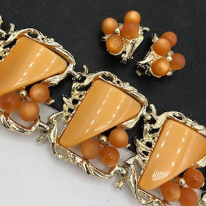May include: A gold tone necklace with a geometric design and orange colored stones. The necklace is paired with matching earrings.