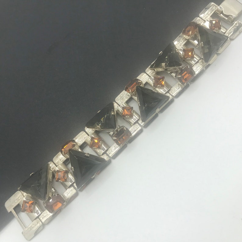 May include: A silver tone bracelet with black and amber coloured rhinestones. The bracelet is made of a series of linked squares with a triangle shaped rhinestone in each square.