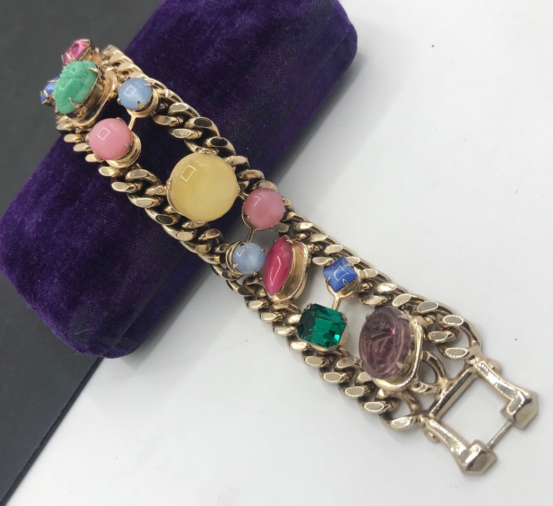 May include: A gold chain bracelet with a double row of colorful stones. The stones are in various shapes and sizes, including round, oval, and square. The stones are set in gold and are surrounded by gold chain links. The bracelet has a clasp closure.
