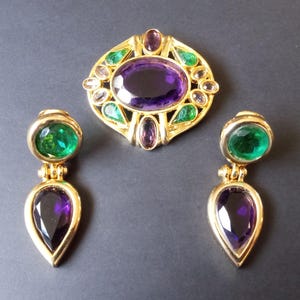 Peut inclure: Une broche et des boucles d'oreilles assorties en or avec des pierres pr&eacute;cieuses vertes et violettes. La broche pr&eacute;sente une grande pierre violette de forme ovale entour&eacute;e de pierres vertes et claires plus petites. Les boucles d'oreilles pr&eacute;sentent une pierre ronde verte et une pierre violette en forme de larme.