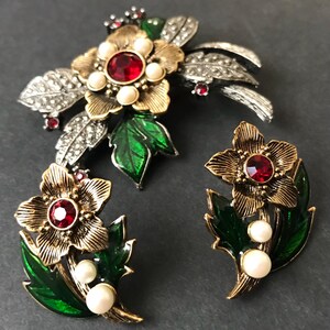Puede incluir: Un conjunto de joyas vintage con un broche floral y pendientes a juego. El broche tiene una flor dorada con una piedra central roja, rodeada de perlas de imitaci&oacute;n y hojas plateadas. Los pendientes reflejan el dise&ntilde;o floral.