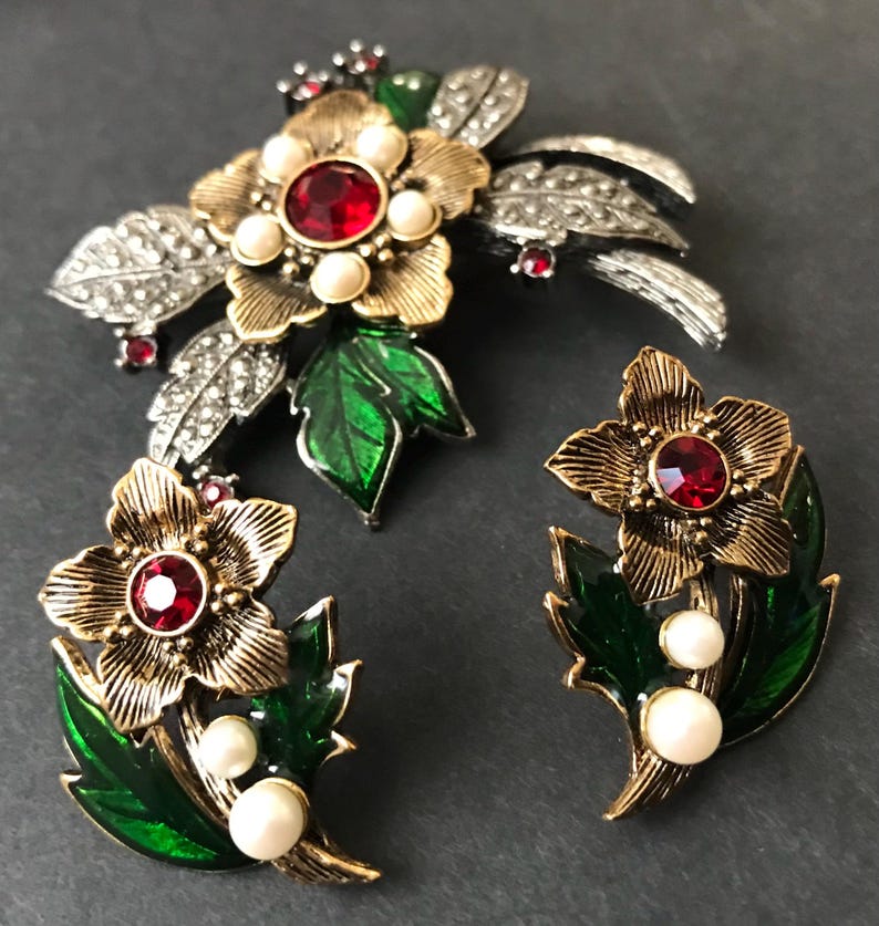 Puede incluir: Un conjunto de joyas vintage con un broche floral y pendientes a juego. El broche tiene una flor dorada con una piedra central roja, rodeada de perlas de imitaci&oacute;n y hojas plateadas. Los pendientes reflejan el dise&ntilde;o floral.