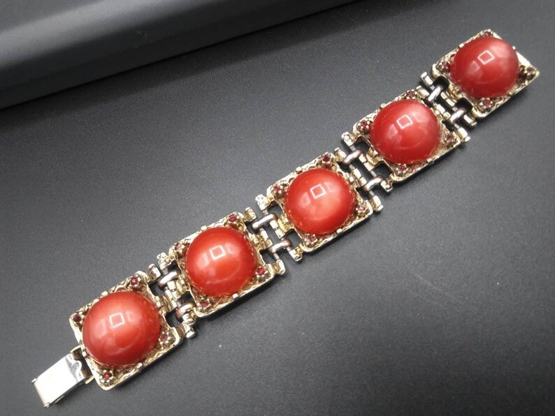 May include: A vintage bracelet featuring five large, round, coral-colored cabochons set in gold-toned metal frames. The bracelet has a silver-toned chain link design and is secured with a clasp. Small red rhinestones accent the frames.
